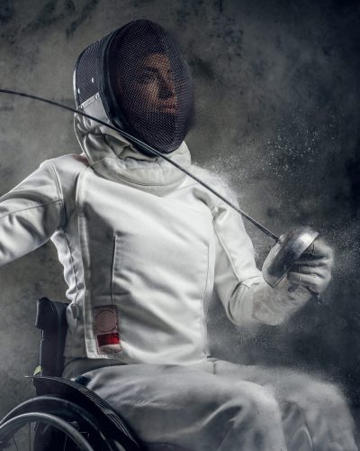 Female fencer in wheelchair with safety mask on a face holding rapier, dust effect on image.